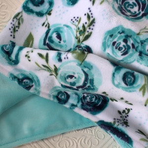 May include: A soft, plush blanket with a white background and a teal floral pattern. The blanket has a light teal minky backing.