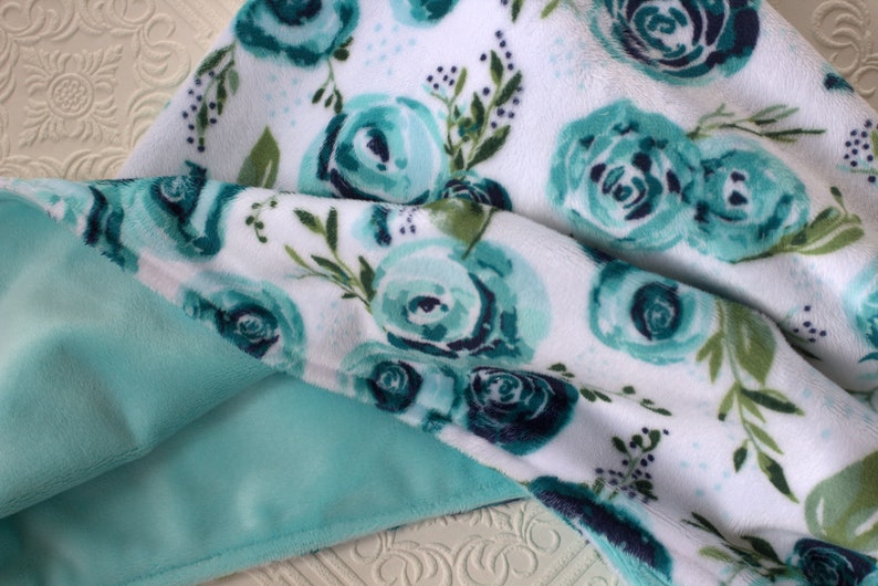 May include: A soft, plush blanket with a white background and a teal floral pattern. The blanket has a light teal minky backing.