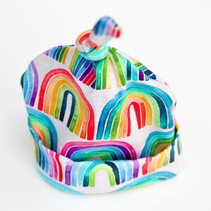 May include: A white baby hat with a rainbow pattern. The hat is tied at the top with a knot.