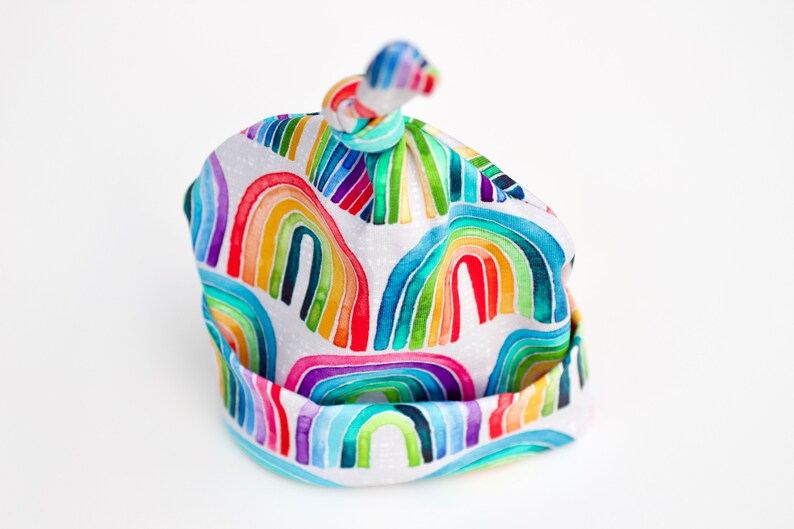 May include: A white baby hat with a rainbow pattern. The hat is tied at the top with a knot.