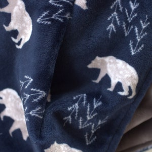 Op de afbeelding: Een marineblauwe fleece deken met een wit beren- en dennenboompatroon. De deken is gevouwen en heeft een zachte, pluche textuur.