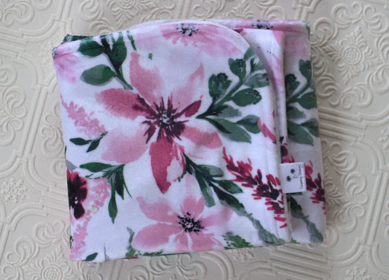 May include: A white baby blanket with a pink and green floral print. The blanket is folded in half and has a small tag that says "Sweet Dreams".