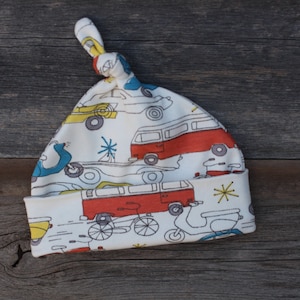 May include: White baby hat with a knot on top. The hat has a pattern of red, yellow, and blue cars, vans, and scooters.
