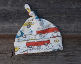 May include: White baby hat with a knot on top. The hat has a pattern of red, yellow, and blue cars, vans, and scooters.