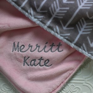 May include: A pink and gray baby blanket with a white arrow pattern on the gray side. The pink side has the name "Merritt Kate" embroidered in gray thread.