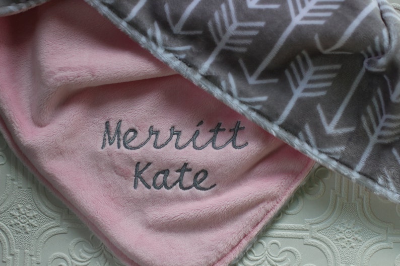 May include: A pink and gray baby blanket with a white arrow pattern on the gray side. The pink side has the name "Merritt Kate" embroidered in gray thread.