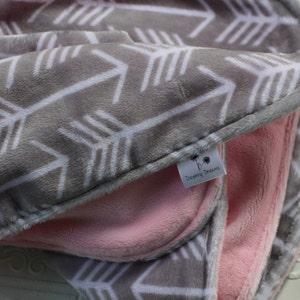 May include: A gray and white arrow print baby blanket with a pink fleece lining. The blanket has a small tag that says "Twirling Dreams".