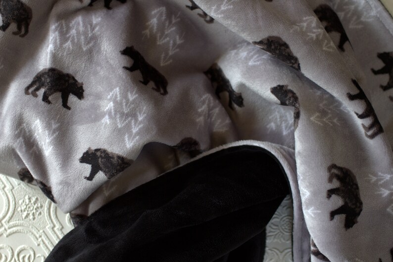 May include: A soft, grey and black fleece blanket with a repeating pattern of bears and trees. The blanket is folded in half, showing both sides.