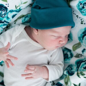May include: A newborn baby wearing a teal blue knitted hat and a white long-sleeved onesie, sleeping on a white blanket with a teal blue floral pattern.