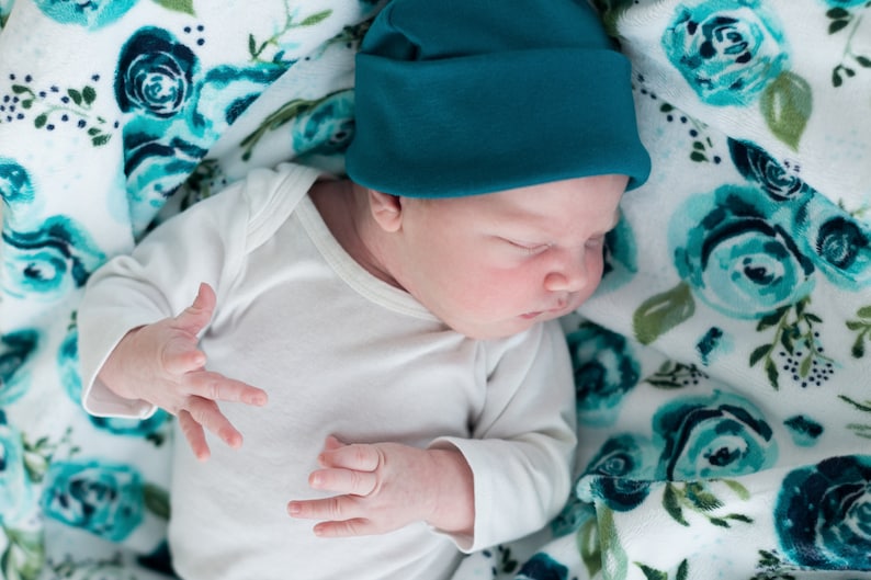 May include: A newborn baby wearing a teal blue knitted hat and a white long-sleeved onesie, sleeping on a white blanket with a teal blue floral pattern.