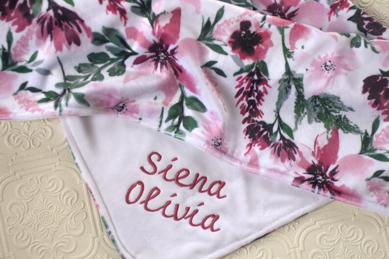May include: White baby blanket with a pink floral print and the names "Siena Olivia" embroidered in pink on the white side.