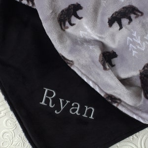 May include: A black fleece blanket with the name "Ryan" embroidered on it. The blanket is folded in half, revealing a grey fleece side with a pattern of black bears.