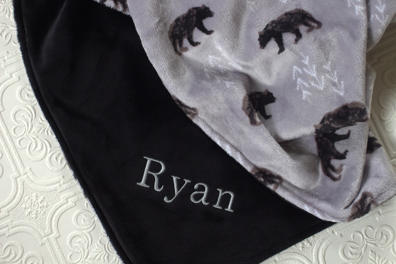 May include: A black fleece blanket with the name "Ryan" embroidered on it. The blanket is folded in half, revealing a grey fleece side with a pattern of black bears.