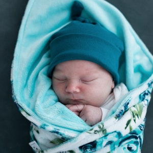 May include: A sleeping baby wrapped in a blue and white floral swaddle blanket with a teal blue knotted beanie on their head.