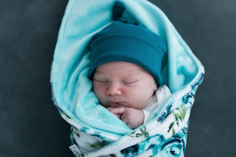 May include: A sleeping baby wrapped in a blue and white floral swaddle blanket with a teal blue knotted beanie on their head.