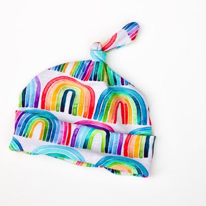 May include: A white baby hat with a rainbow pattern. The hat has a knot on top and is made of soft fabric.