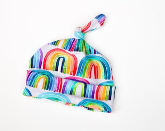 May include: A white baby hat with a rainbow pattern. The hat has a knot on top and is made of soft fabric.