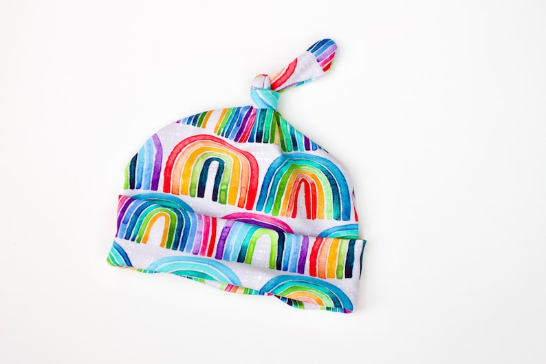 May include: A white baby hat with a rainbow pattern. The hat has a knot on top and is made of soft fabric.