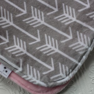 May include: A gray and white baby blanket with a chevron pattern of white arrows on a gray background. The blanket has a pink fleece backing and a white tag with the text "Dreaming Dreams".