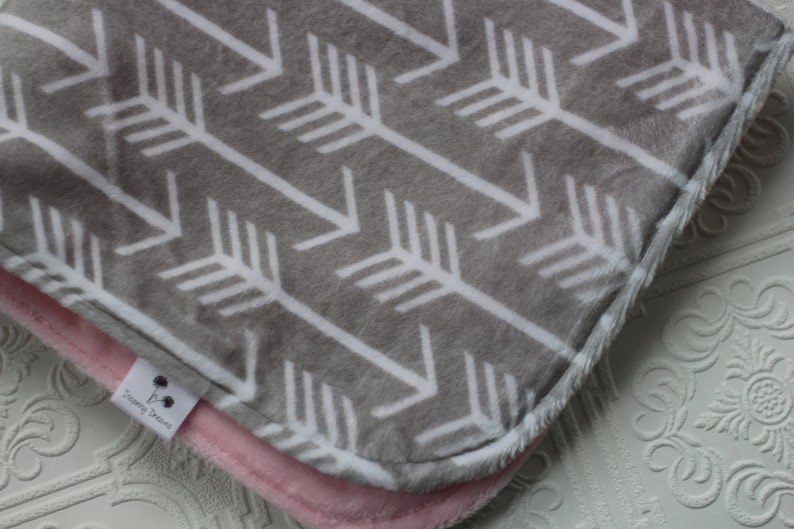 May include: A gray and white baby blanket with a chevron pattern of white arrows on a gray background. The blanket has a pink fleece backing and a white tag with the text "Dreaming Dreams".