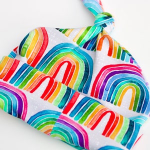 May include: A white baby hat with a rainbow pattern. The hat is tied at the top with a knot.