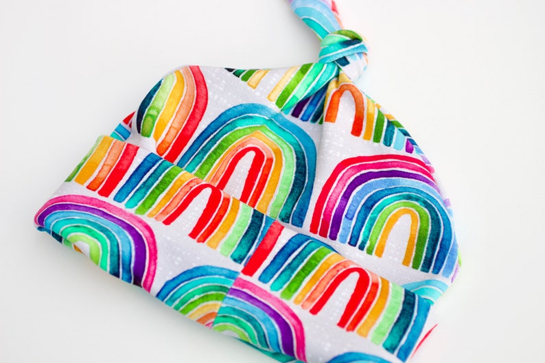 May include: A white baby hat with a rainbow pattern. The hat is tied at the top with a knot.