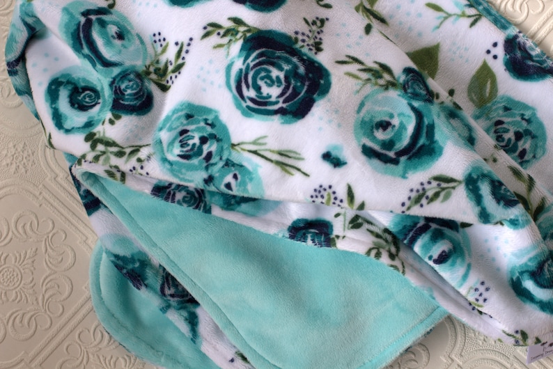 May include: A soft, plush throw blanket with a white background and a teal floral pattern. The blanket has a light teal underside.