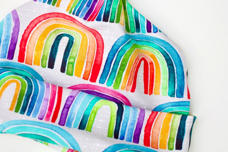 May include: A white beanie hat with a colorful rainbow pattern. The rainbow is made up of watercolor-like stripes in shades of red, orange, yellow, green, blue, indigo, and violet.