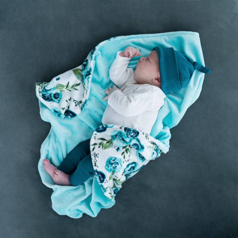 May include: A baby wearing a teal blue hat and white onesie is sleeping on a turquoise blue and white floral blanket.
