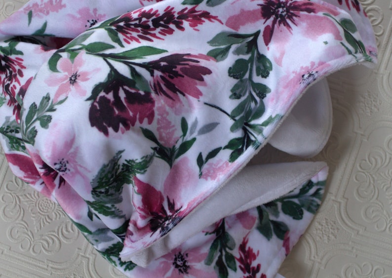 May include: A white blanket with a floral pattern in shades of pink and green. The blanket is folded in half and the fabric is soft and plush.