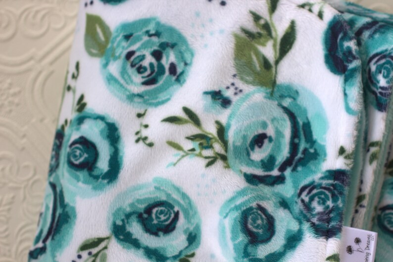 May include: White plush blanket with a teal floral pattern. The blanket features a repeating pattern of teal roses with green leaves.