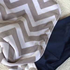 May include: A soft, plush blanket with a chevron pattern in beige and white. The blanket is folded in half and partially covers a dark blue blanket.
