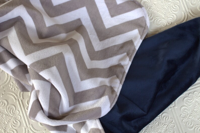 May include: A soft, plush blanket with a chevron pattern in beige and white. The blanket is folded in half and partially covers a dark blue blanket.
