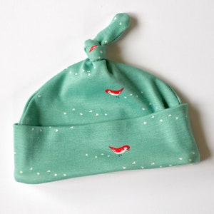 May include: A green baby hat with a white polka dot pattern and red birds. The hat has a knot on top.
