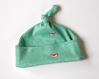 May include: A green baby hat with a white polka dot pattern and red birds. The hat has a knot on top.