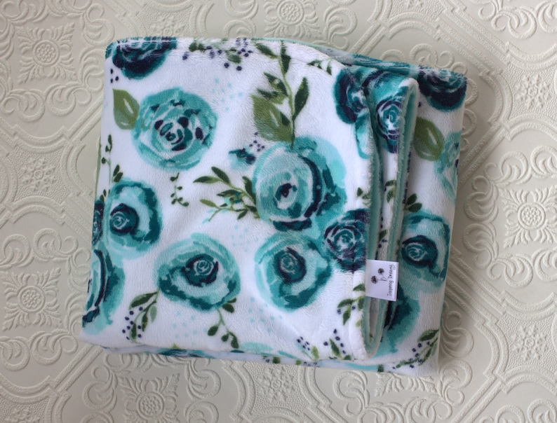 May include: A white and teal floral print blanket with a soft, plush texture. The blanket is folded in half and has a small tag that says "Sleeping Dreams".