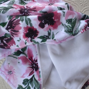May include: A white fabric baby blanket with a pink and green floral print. One side of the blanket is soft and fuzzy, while the other side is smooth and silky. The blanket has a small tag that says "Dreaming Dreams".