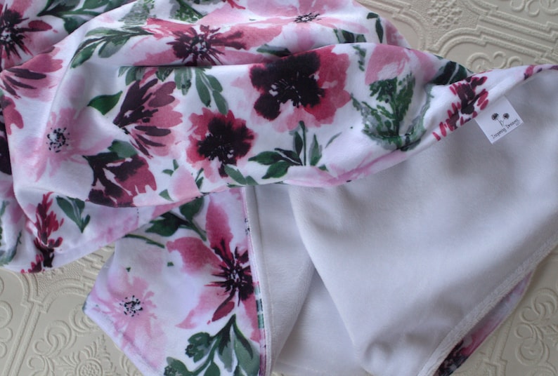 May include: A white fabric baby blanket with a pink and green floral print. One side of the blanket is soft and fuzzy, while the other side is smooth and silky. The blanket has a small tag that says "Dreaming Dreams".