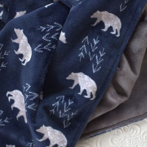 Op de afbeelding: Een donkerblauwe fleece deken met een wit beren- en dennenboompatroon. De deken is dubbelgevouwen en de andere kant is effen grijs fleece.