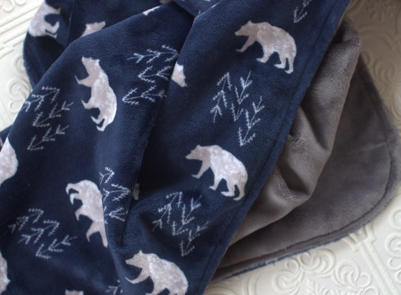 Op de afbeelding: Een donkerblauwe fleece deken met een wit beren- en dennenboompatroon. De deken is dubbelgevouwen en de andere kant is effen grijs fleece.