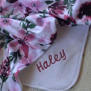 May include: A white baby blanket with a floral print in shades of pink and green. The blanket has a white border and the name "Haley" is embroidered in pink on the white border.