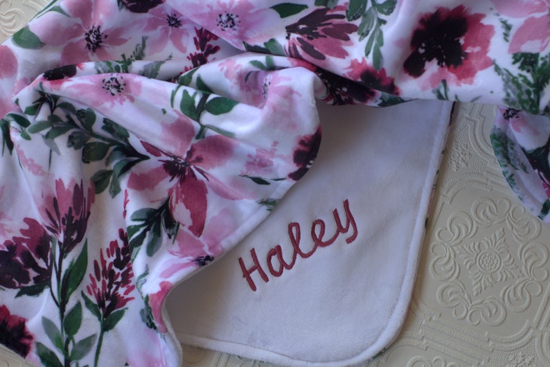 May include: A white baby blanket with a floral print in shades of pink and green. The blanket has a white border and the name "Haley" is embroidered in pink on the white border.