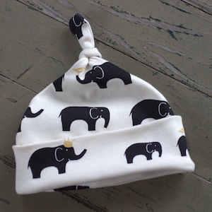 May include: White baby hat with a black elephant print. The hat has a knot on top and a gold crown on one of the elephants.