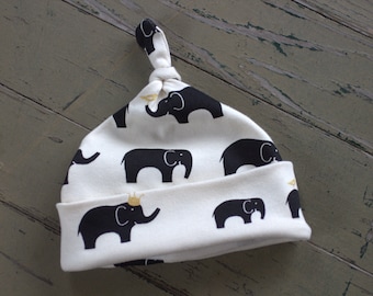 May include: White baby hat with a black elephant print. The hat has a knot on top and a gold crown on one of the elephants.