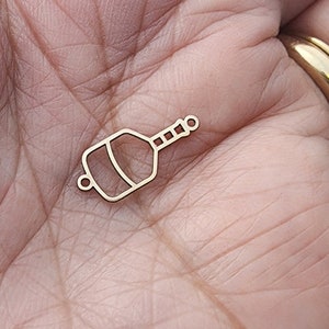 May include: A gold-colored charm in the shape of a champagne bottle. The charm has a rectangular body with a rounded bottom and a long, thin neck with a small circle at the end. The charm is made of a thin metal.