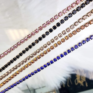 May include: Five rows of rhinestone chain trim in different colors, including pink, black, brown, orange, and blue. The trim is made of small square rhinestones connected by gold metal.