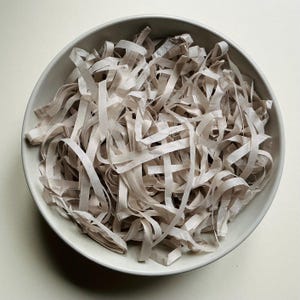 May include: A round, light-colored ceramic bowl filled with a generous amount of thin, light beige paper strips. The paper is cut into long, narrow ribbons that are loosely piled and tangled together, creating a textured, layered appearance.