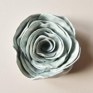 Pode incluir: Escultura de rosa em cerâmica, num tom verde-azulado suave e discreto. A peça artesanal apresenta pétalas em camadas que se enrolam para dentro, criando uma forma floral realista. Um elemento decorativo para a decoração da casa.