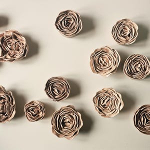 Peut inclure: Une collection de décorations murales en forme de roses beiges faites à la main. Les roses varient en taille et sont disposées de manière éparse sur un fond neutre. Les pétales détaillés créent un design floral réaliste, adapté à la décoration intérieure.