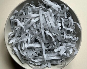 May include: A round, light gray ceramic bowl filled with a large quantity of thin, light gray paper strips. The paper strips are arranged in a tangled, layered pile, completely covering the interior base of the bowl.